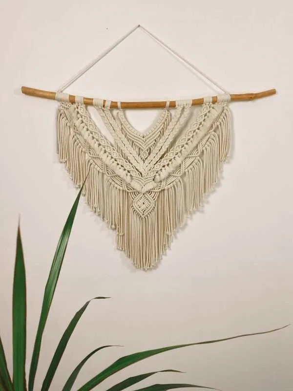 Painel de macramé grande Savanna com tranças em V e franja em ponta, ao lado de uma planta