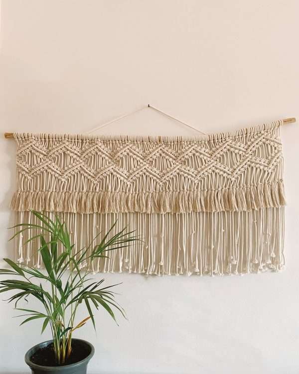Painel de macramé horizontal Horizon com losangos e franja dupla, por cima de uma palmeira em vaso