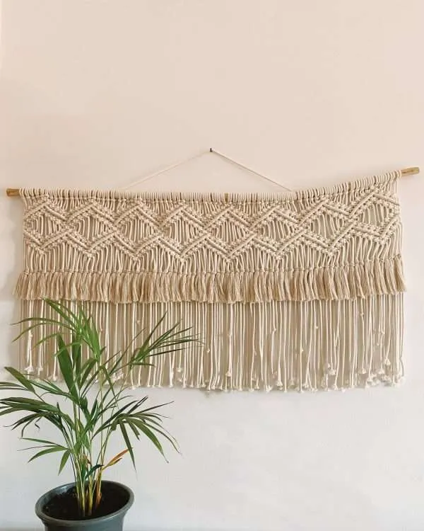 Painel de macramé horizontal Horizon com losangos e franja dupla, por cima de uma palmeira em vaso