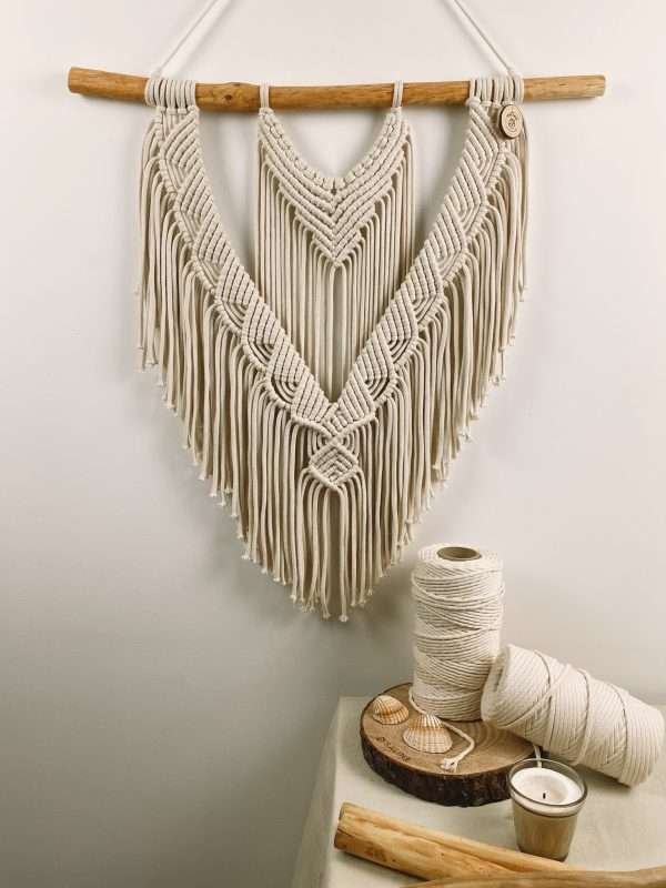 Painel de parede em macramé Coral com linhas em V e franja em ponta, ao lado de rolos de algodão