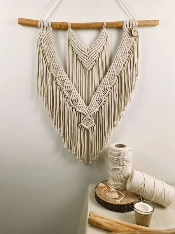 Painel de parede em macramé Coral com linhas em V e franja em ponta, ao lado de rolos de algodão