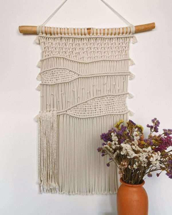 Painel de parede em macramé Roots com faixas onduladas e franja solta, ao lado de flores secas