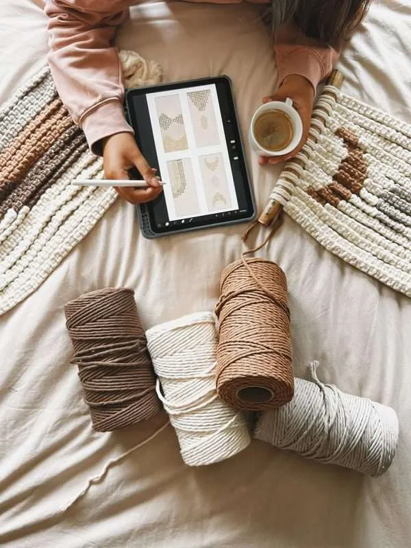 Desenhos de peças personalizadas num tablet, ao lado de rolos de fio de algodão e painéis de macramé em tons de terra