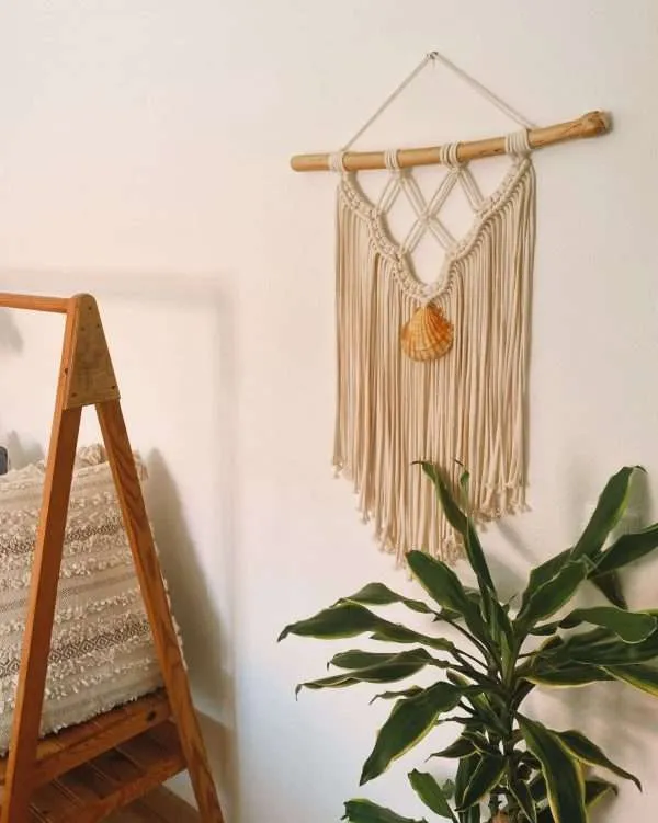 Painel de parede em macramé Ocean com concha ao centro, ao lado de um espelho e de uma planta