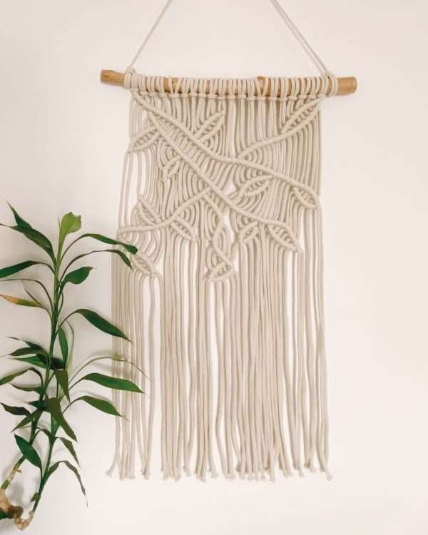 Painel de parede em macramé Forest com ramos e folhas em diagonal, ao lado de uma planta