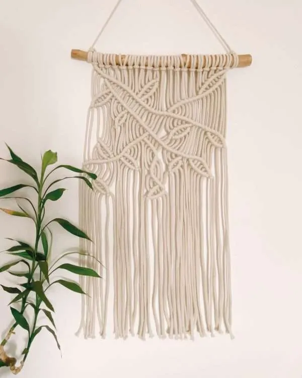 Painel de parede em macramé Forest com ramos e folhas em diagonal, ao lado de uma planta